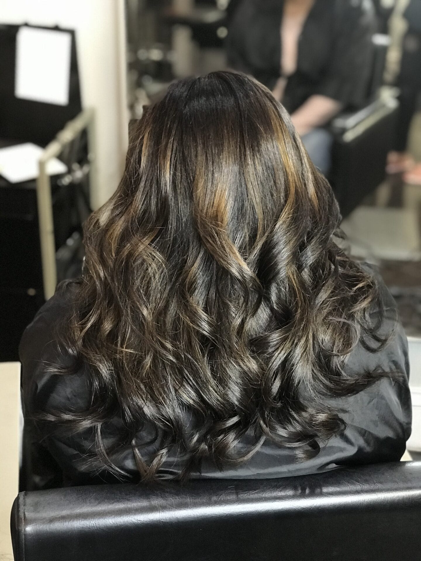 A person with long, wavy, dark brown hair and caramel highlights sits in a salon chair at a San Francisco hair salon, showcasing expert hair color & lighting while facing away from the camera.