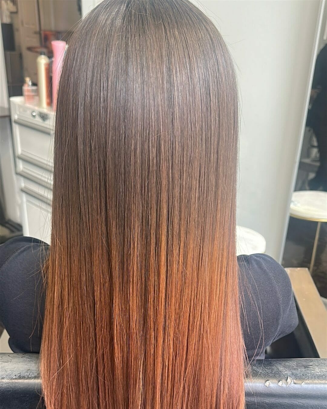 Person with straight, long hair in a brown to copper ombre style, fresh from a Brazilian Blowout, sitting in a salon chair.