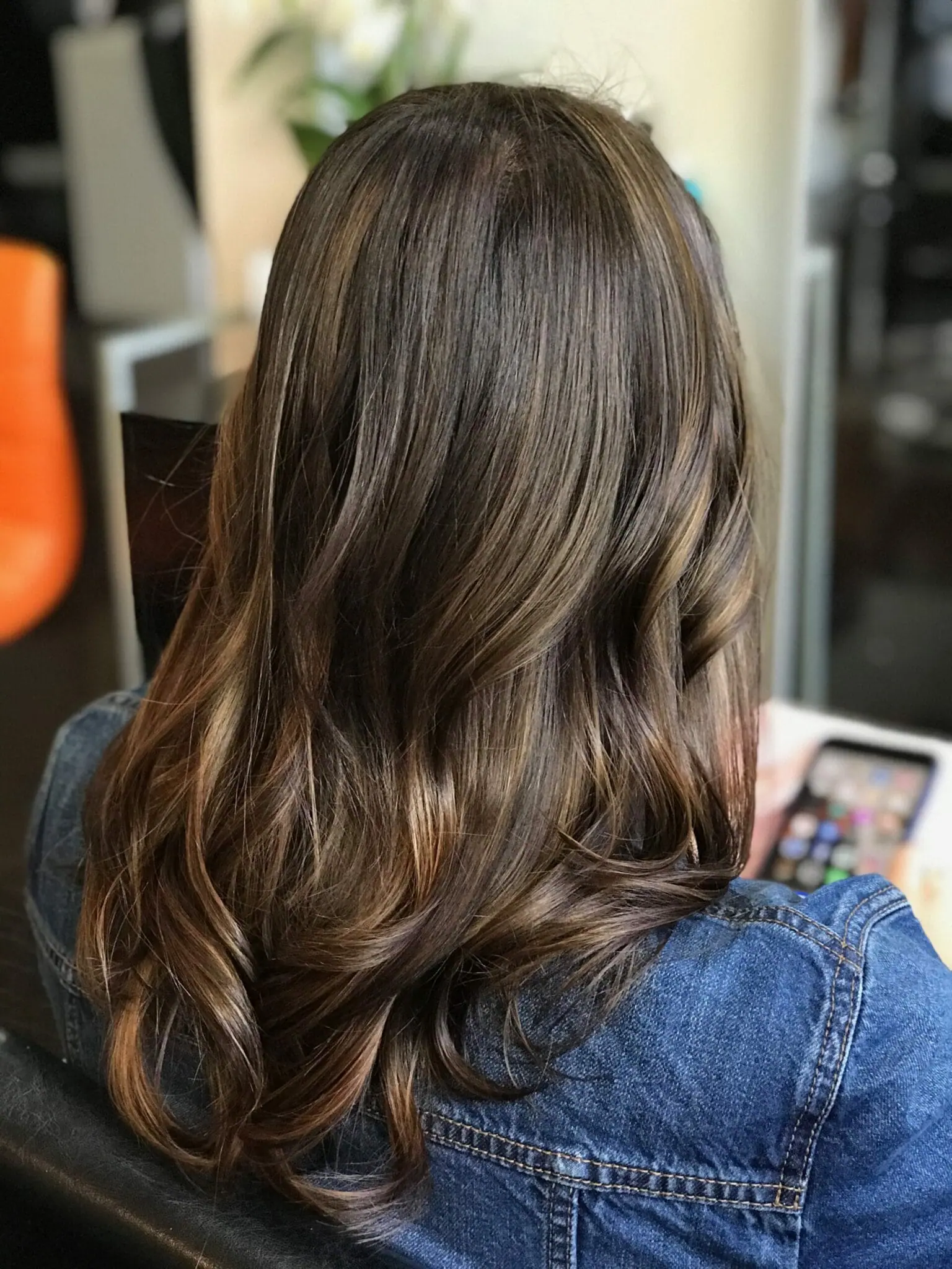 Woman with brown wavy hair wearing a denim jacket, seated and holding a smartphone.